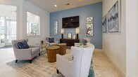 Located at 1301 Charter Colony Pkwy in Midlothian, Virginia, this thoughtfully designed single-floor amenity space offers a welcoming and upscale environment for gathering and socializing. The open-concept layout flows seamlessly between several distinct functional areas, all unified by elegant marble-look tile flooring that extends throughout.

The centerpiece of the space is a well-appointed kitchen featuring a large island with a bold blue countertop and seating for multiple guests, sleek taupe cabinetry, stainless steel appliances, and a striking slatted ceiling detail with pendant lighting overhead. The kitchen opens directly into a spacious communal dining area lined with upholstered chairs, teal drapery, and ample natural light streaming through large windows.

Adjacent to the dining area, a bright and airy seating nook features a round marble-top table surrounded by teal velvet accent chairs with nailhead trim, positioned beside French doors that open to an outdoor terrace with views of the surrounding community.

A cozy lounge area anchors another corner of the space, highlighted by a dusty blue accent wall, a wall-mounted television, and an arrangement of plush gray upholstered seating centered around warm wood accent tables. Curated artwork and decorative accents throughout lend a polished, cohesive aesthetic to the entire space.