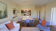 This single-floor residence offers a thoughtfully designed open-concept layout that seamlessly connects the living, dining, and kitchen areas. The dining space is anchored by a rectangular white table surrounded by contemporary gray chairs, positioned directly in front of a pass-through window that opens to the kitchen — a practical and stylish design choice that enhances the sense of openness. The kitchen is equipped with stainless steel appliances, including a refrigerator, microwave, and range, complemented by dark cabinetry and warm pendant lighting that adds a refined touch overhead. Track lighting in the main living and dining area provides even, adjustable illumination throughout the space. Rich, dark hardwood flooring runs throughout, grounding the interior with warmth and continuity, while light-toned area rugs define the living zone and add texture. The living area features clean-lined white seating accented with coral throw pillows, creating a inviting, casual atmosphere. A decorative mirror and greenery on a wall-mounted shelf add personality to the space. Neutral wall tones keep the interior feeling bright and airy. A hallway visible in the background suggests additional private rooms beyond, completing a functional and cohesive residential layout.