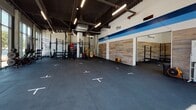 Located at 435 Melnea Cass Blvd in Boston, MA, this single-floor fitness facility offers a well-designed, open-concept training environment in the Roxbury neighborhood. The space is defined by its expansive floor-to-ceiling glass curtain wall along the front façade, flooding the interior with abundant natural light and providing clear views of the surrounding streetscape and greenery outside.

The main training floor is generously proportioned, featuring durable rubber flooring with marked station indicators, allowing for organized group or individual workouts. Soaring ceilings with exposed industrial-style ductwork and overhead linear lighting give the space an open, energetic atmosphere. A bold blue horizontal stripe and branded signage accent the white walls, reinforcing the facility's identity.

Along the perimeter, wood-paneled accent walls add warmth and texture, complementing the otherwise industrial aesthetic. Equipment such as squat racks, pull-up rigs, medicine balls, weight plates, and stationary bikes are positioned throughout, supporting a variety of training modalities. A whiteboard station provides a functional coaching area. The glass entry vestibule offers a clean, welcoming transition from the outdoors into the facility.