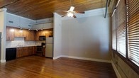 This single-floor loft-style space blends industrial character with warm, inviting finishes throughout. The open layout is anchored by rich, dark hardwood floors that run seamlessly from the living area into the kitchen, creating a cohesive and flowing interior.

The ceiling is a standout architectural feature, finished with natural wood planks that add texture and warmth to the space. A ceiling fan with a built-in light fixture is centrally positioned, providing both air circulation and ambient lighting.

The kitchen is well-appointed and functional, featuring stainless steel appliances including a refrigerator, electric range, and built-in microwave. Warm wood cabinetry lines the walls, complemented by a classic tile backsplash and under-cabinet track lighting that illuminates the workspace. The kitchen layout makes efficient use of the available space while maintaining an open connection to the rest of the interior.

Exposed industrial elements, including visible ductwork or piping along the upper walls, add an authentic loft aesthetic that contrasts beautifully with the natural wood tones. The neutral-toned walls provide a versatile backdrop, allowing the architectural details to take center stage. Overall, the space offers a distinctive character that balances rustic warmth with modern functionality.