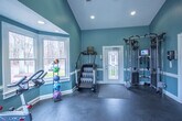 Located at 101 Little Bay Ave in Yorktown, Virginia, this dedicated fitness facility offers a thoughtfully designed single-floor workout space that combines functionality with an inviting atmosphere. The room features vaulted ceilings with recessed lighting and a ceiling fan, creating an open, airy feel throughout.

The walls are finished in a calming blue-gray tone, complemented by white chair-rail molding that adds a classic architectural detail. Durable dark rubber flooring covers the entire space, providing both safety and practicality for exercise use. Multiple windows along the walls, including a charming bay window, flood the room with natural light and offer views of the surrounding wooded landscape.

The gym is comprehensively equipped for a full-body workout. Cardio offerings include two treadmills, an elliptical trainer, a stair climber, and a stationary spin bike. Strength training is well-supported by a multi-functional cable machine system, a medicine ball rack, free weights, and additional resistance equipment. A water cooler is conveniently positioned within the space. A decorative glass-panel entry door provides a welcoming point of access. Overall, this fitness space delivers a professional-grade workout environment within a well-appointed, naturally lit setting.