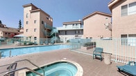 This residential community showcases a warm, Southern California-inspired architectural style, characterized by earth-toned stucco exteriors, tiled rooflines, and decorative arched details that lend the complex a cohesive and inviting aesthetic. The centerpiece of the outdoor common area is a generously sized swimming pool with crystal-clear turquoise water, complemented by a separate bubbling spa for relaxation. The pool deck is finished in warm terracotta-toned tile, offering ample space for lounge chairs and outdoor furniture, perfect for soaking up the abundant sunshine. A teal-painted metal fence encloses the pool area, providing safety while maintaining an open, airy feel. Lush green landscaping and umbrella-shaded seating areas add pockets of shade and greenery throughout the space. The surrounding multi-story buildings feature private balconies, exterior staircases in a matching teal accent color, and a mix of traditional and contemporary window designs. An arched passageway detail on one of the buildings adds a distinctive architectural touch. The complex benefits from clear open skies and a bright, sun-drenched atmosphere, creating an enjoyable outdoor living environment for residents throughout the year.