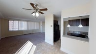 This single-floor residence offers an open and functional layout that seamlessly connects the living area and kitchen. The living room is generously sized, featuring soft carpeted flooring, a ceiling fan with integrated lighting, and a large window dressed with vertical blinds that floods the space with natural light. A baseboard radiator runs along the windowed wall, providing efficient heating throughout the area.

The kitchen is thoughtfully positioned adjacent to the living space, separated by a structural column that adds architectural definition. It features dark espresso-toned cabinetry, a black refrigerator, and a dark range, all set against stone-patterned countertops. A pass-through counter with a pendant light above creates an open connection between the kitchen and the main living area, enhancing the sense of flow. The kitchen is finished with the same tile flooring found in the entryway, providing a practical and cohesive transition from the main living space.

Overall, the residence presents a clean, neutral palette of beige and white tones throughout, lending a bright and welcoming ambiance. The layout is efficient and well-organized, making excellent use of the available footprint while maintaining a comfortable and livable atmosphere.