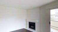 This single-floor space presents a clean and minimalist aesthetic, defined by smooth, neutral-toned walls and bright, open interiors. The living area serves as a central focal point, anchored by a built-in fireplace with a crisp white surround and mantel. The fireplace adds both architectural character and a warm, inviting ambiance to the room. The flooring transitions between classic white tile near the hearth and rich dark hardwood, creating a subtle yet visually appealing contrast throughout the space. The ceilings are generously proportioned, contributing to an airy and expansive feel. The walls are finished in a soft, off-white palette that reflects light evenly, enhancing the brightness of the interior. Electrical outlets are thoughtfully positioned throughout the room, offering practical functionality. A fire suppression sprinkler is visible on the ceiling, indicating that the space meets modern safety standards. Overall, the space offers a versatile and well-maintained environment with a simple, timeless design that provides an excellent foundation for a variety of interior styles and uses.