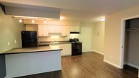 Located at 5400 Portree Pl in Raleigh, NC, this single-floor residence offers a thoughtfully designed open-concept layout that seamlessly connects the kitchen and living areas. The kitchen is well-appointed with white cabinetry providing ample storage, dark countertops, and a full suite of black appliances including a refrigerator, electric range, and built-in microwave. A peninsula countertop extends into the living space, creating a natural divide between cooking and relaxing areas while maintaining an open, airy feel. Under-cabinet lighting illuminates the workspace, complementing the recessed and track lighting overhead. Rich, dark hardwood-style flooring runs throughout the main living space, adding warmth and a cohesive visual flow. Neutral-toned walls keep the interior feeling bright and versatile, ready to accommodate a variety of décor styles. A paneled interior door leads to additional areas of the unit, and clean white baseboards frame the space with a crisp, finished look. The overall design balances functionality and comfort, making efficient use of the single-level floor plan.