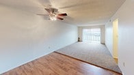 This single-floor unit offers a practical and functional layout with clearly defined living spaces. The galley-style kitchen is lined on both sides with warm honey-toned wood cabinetry, offering generous upper and lower storage. Light-toned countertops and a white electric range complete the space, which opens directly into the main living and dining area.

The living area features a combination of wood-look hard flooring and plush carpeting, creating distinct zones within the open layout. A ceiling fan with a light fixture provides both ventilation and overhead lighting. A large sliding glass door fitted with vertical blinds allows natural light to fill the room and provides access to the exterior.

Adjacent to the living space, the bedroom is carpeted and features neutral walls, a textured ceiling, and a window with horizontal blinds that admits natural light. The room offers a quiet, functional setting suitable for a variety of furniture arrangements.

The bathroom is efficiently laid out with wood-look flooring, a full-size soaking tub, and a tiled shower surround featuring large-format neutral tiles with a contrasting dark accent strip. A wall-mounted towel bar and overhead lighting complete the space. Throughout the unit, textured ceilings and a neutral color palette provide a consistent, understated aesthetic.