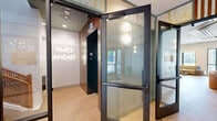 Located at 55 South St in South Walpole, MA, this thoughtfully designed two-floor amenity space offers an impressive blend of functionality and curated interior design across a generous footprint. The ground floor welcomes residents through a stylish entry corridor finished with teal-toned paneling, warm wood-slat ceiling accents, and wall sconces, setting an immediate tone of sophistication. Adjacent to the entry, a well-appointed fitness center commands double-height ceilings with a vibrant, large-scale mural as its focal point, while an assortment of cardio machines, strength equipment, and free weights occupy a boldly patterned tile floor. A dedicated exercise room with direct outdoor access rounds out the wellness offerings. The upper floor transitions into a dynamic social hub, anchored by a spacious lounge featuring a linear gas fireplace, built-in shelving with curated décor, and a dramatic ceiling adorned with shibori-inspired artwork framed by warm wood beams. Natural light pours in through expansive windows with views of the surrounding landscape. The adjacent kitchen and dining area showcases a striking marble-topped island with bar seating, rich walnut cabinetry, geometric tile backsplash, and premium stainless appliances. A sculptural staircase with an artistic shibori-mural surround and a cascading pendant light connects both floors, while additional hallways and breakout rooms provide versatile spaces throughout.