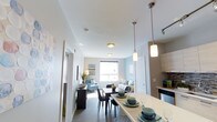 This single-floor residence offers a well-designed open-concept layout that integrates the kitchen, dining, and living spaces into one cohesive and functional environment. The kitchen is positioned at the front, featuring light wood cabinetry, a sleek countertop island with bar seating, and stylish pendant lighting that adds a contemporary touch. The dining area flows naturally from the kitchen, centered around a dark wood table with seating for multiple guests, illuminated by matching pendant fixtures overhead. Beyond the dining space, the living room is furnished with comfortable seating in soft blue tones, complemented by light grey walls that create a calm and welcoming atmosphere. A window at the far end of the living area allows natural light to fill the space, enhancing the airy and open feel. Hardwood-style flooring runs throughout, adding warmth and visual continuity across all areas. Recessed and track lighting provide additional illumination, ensuring the space is well-lit at all times. A corridor leading off the main living area suggests additional rooms, offering privacy and separation from the communal spaces. Overall, the residence presents a clean, modern aesthetic with a thoughtfully designed layout suited for everyday living.