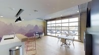 This contemporary single-floor space is thoughtfully designed to foster collaboration and creativity. The interior is immediately distinguished by a large-scale wall mural depicting a serene, layered mountain landscape in soft purples and warm neutrals, adding an artistic focal point to the room.

Natural light floods the space through an expansive floor-to-ceiling garage-style glass door, which spans nearly an entire wall and provides sweeping views of the outdoor surroundings. The industrial-style door hardware complements the exposed concrete ceiling elements, lending an urban, loft-like character to the environment.

Light hardwood flooring runs throughout, contributing warmth and cohesion to the open layout. The space is furnished with multiple seating arrangements to accommodate a variety of uses. A round black bistro table paired with white chairs sits near the mural, while a larger oval white table surrounded by modern white shell chairs is positioned closer to the glass wall, ideal for group meetings or casual gatherings. A counter-height bar along one side features distinctive pink wire bar stools, adding a playful pop of color. Recessed lighting in the white ceiling supplements the abundant natural light, ensuring the space remains bright and welcoming at all times.