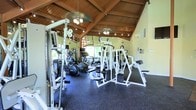 This fitness center offers a thoughtfully designed workout environment that combines architectural character with practical functionality. The space is defined by its striking vaulted ceiling with exposed wood beams, which lends a warm, lodge-inspired aesthetic uncommon in typical gym settings. Ceiling fans provide comfortable air circulation throughout the workout area, while track lighting ensures consistent, well-distributed illumination across the floor.

The rubber-floored workout area is outfitted with a robust selection of strength training machines, including multi-station cable and weight machines in white frames, a flat bench press setup, and a squat rack. On the cardio side, the space features elliptical trainers and treadmills positioned near large windows that allow natural light to flow in and provide outdoor views, creating a more engaging exercise environment.

Full-length mirrored walls line the space, visually expanding the room and allowing users to monitor their form during workouts. The combination of the high vaulted ceiling, mirrored walls, and natural light contributes to an open and airy atmosphere. The overall layout is organized to facilitate both cardio and strength training routines, making this a versatile, well-rounded fitness facility suitable for a wide range of exercise needs.