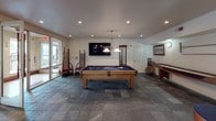This single-floor space is thoughtfully designed for entertaining and recreation, combining a well-appointed kitchen with a generous game room area. The kitchen is outfitted with rich, warm-toned wood cabinetry that extends floor to ceiling, providing ample storage throughout. Light granite countertops offer a striking contrast against the cabinetry, while a diamond-patterned tile backsplash with under-cabinet LED lighting adds a refined touch. Stainless steel appliances — including a double wall oven, refrigerator with ice maker, dishwasher, and built-in microwave — equip the kitchen for full-scale entertaining. A central island with a cooktop and granite surface provides additional workspace. The flooring throughout is durable slate tile in a muted, earthy tone that ties the entire space together seamlessly. The expansive game room is the centerpiece of the space, anchored by a full-size pool table with a pendant light overhead and a wall-mounted cue rack nearby. A large flat-screen TV is mounted prominently on the wall, complemented by recessed lighting and circular wall sconces. A shuffleboard table and bar-height seating add further entertainment options. Sliding glass doors open to an outdoor area, allowing natural light to fill the room and creating an easy indoor-outdoor flow.