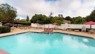 Located at 715 Rice Rd in Ridgeland, Mississippi, this single-level outdoor amenity space centers around a generously sized swimming pool with crystal-clear blue water. A distinctive brick fountain feature sits at the heart of the pool, providing an attractive focal point and the gentle sound of flowing water. The pool is framed by a wide concrete and aggregate deck that offers ample room for relaxation, furnished with multiple lounge chairs and seating arrangements shaded by vibrant red patio umbrellas. A brick pool house structure with a pitched roof sits adjacent to the pool area, providing a functional support facility for the space. The entire area is enclosed by a white vinyl privacy fence, creating a secure and intimate environment. Lush mature trees and manicured hedges line the perimeter, offering natural shade and a verdant backdrop that enhances the overall atmosphere. The combination of classic brick architecture, well-maintained landscaping, and thoughtful amenities makes this outdoor pool area a welcoming and practical community gathering space.