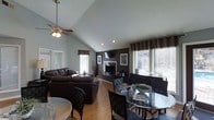 This single-floor home showcases a thoughtfully designed open-concept layout that seamlessly connects the living and dining areas into one cohesive, welcoming space. The vaulted ceiling rises dramatically overhead, adding a strong sense of volume and airiness, complemented by a ceiling fan that enhances both comfort and visual interest.

The living area is anchored by a striking dark accent wall housing a fireplace and a mounted television, creating a bold focal point that contrasts beautifully with the lighter tones of the surrounding walls and smooth hardwood-style flooring. Rich, dark leather sofas furnish the space with a sense of warmth and sophistication.

The dining area sits adjacent to the living room, featuring a glass-topped table with dark woven chairs, offering an elegant yet casual setting for meals. Multiple large windows dressed with dark curtains and horizontal blinds flood the interior with natural light while framing views of the outdoors.

Throughout the space, the interplay of dark furnishings against lighter walls and flooring creates a balanced, inviting atmosphere. Recessed lighting and track lighting further illuminate the area, making it functional and well-lit at any time of day. The overall design reflects a modern, comfortable aesthetic suited for everyday living and entertaining alike.