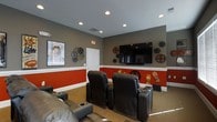 This single-story screening room is designed with an unmistakable cinematic aesthetic. The walls are finished in a two-tone scheme, with warm red on the lower half separated by crisp white wainscoting and a cool gray on the upper half, creating a classic movie house atmosphere. Recessed lighting in the gray ceiling provides ambient illumination throughout the space.

The focal point of the room is a large wall-mounted flat-screen television, positioned front and center for optimal viewing from all seating positions. Multiple rows of black leather recliner-style chairs, each equipped with built-in cup holders, provide comfortable, theater-style seating arrangements. The carpeted flooring adds warmth and aids in sound absorption.

Decorative film reel wall art is displayed throughout the room, reinforcing the cinema theme, alongside a framed movie poster. Additional themed décor pieces, including figurines and metallic reel accents in varying sizes, are arranged across the walls. A standard door provides access to the rest of the space. The overall ambiance is purposefully immersive, designed to replicate the feel of a traditional movie theater in a private setting.