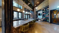 This thoughtfully designed amenity space makes an immediate impression with its soaring double-height ceilings and bold, eclectic interiors. The centerpiece of the main area is a sweeping granite-topped bar island, accented by elegant globe pendant lights suspended from brass fixtures and surrounded by sculptural green velvet bar stools with gold-toned legs. Behind the bar, a dramatic floor-to-ceiling feature wall clad in richly textured, dark-toned reclaimed wood adds warmth and visual depth. Large black-framed windows along the perimeter flood the space with natural light while offering views of the surrounding outdoor areas. An upper mezzanine level, finished with deep green tile and brass railings, overlooks the main floor below. Adjacent to the bar, a comfortable lounge area offers a relaxed retreat, complete with floor-to-ceiling navy built-in bookshelves, a wall-mounted television, a wooden bench, and plush seating arranged over a decorative area rug. Polished concrete floors run throughout, lending an industrial-chic quality to the space. Corridors connect additional functional areas, including a wet bar prep zone and curated hallways adorned with geometric wood wall art. The overall aesthetic blends contemporary design with organic, textural elements for a refined yet inviting atmosphere.