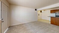 Located at 5201 East 2nd Street in Tucson, Arizona, this one-bedroom, one-bathroom unit offers a practical and efficient single-floor layout. The open living area features neutral carpet flooring and painted brick walls, creating a clean, understated backdrop. A ceiling fan overhead provides airflow and lighting, while the space flows directly into the kitchen area, which is set apart by tile flooring. The kitchen is equipped with warm-toned wood cabinetry — both upper and lower — along with a white refrigerator, electric range, and a double sink with a dark countertop. A recessed light illuminates the workspace, and a peninsula-style counter adds a subtle division between kitchen and living areas. The bedroom is a comfortable, carpeted room featuring painted brick accent walls and windows that bring in natural light. A set of double-door closets provides ample storage, and a separate door connects conveniently to the hallway. The bathroom includes a full tub and shower combination with classic tile surround featuring a decorative border, complemented by a vanity with a light countertop and tile flooring. A hallway connects all rooms, completing this efficient and functional layout.