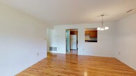 Located at Whiton Hills, 708 Abby, this single-floor residence offers a clean, open layout with a bright and understated aesthetic. The main living area is finished with warm-toned hardwood flooring that extends throughout, reflecting light and contributing to the spacious feel of the interior. The white walls provide a neutral canvas, keeping the space feeling open and airy.

A chandelier with a brushed nickel finish hangs near the kitchen pass-through, adding a subtle decorative touch to the living area. The kitchen features warm honey-toned wood cabinetry, a white refrigerator, and a dark countertop cooktop surface, all visible through the open pass-through window. The kitchen connects to an adjacent utility or laundry area accessible through a doorway, enhancing the functional layout of the unit.

Floor vents are integrated along the baseboards, indicating a forced-air climate system. The overall design is practical and well-maintained, with smooth ceilings and clean trim work throughout. The open connection between the living space and kitchen promotes a cohesive, easy-to-navigate floor plan suited for everyday living.