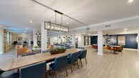 Located at 5391 W Fayetteville Rd in College Park, Georgia, this single-floor community clubhouse offers a spacious and thoughtfully designed gathering space ideal for socializing, working, and relaxing. The open-concept layout is anchored by light hardwood-style flooring, white structural columns, and recessed lighting throughout, creating a bright and airy atmosphere.

The central social area features a large communal dining table with bold blue upholstered chairs beneath an industrial-style rectangular chandelier, complemented by multiple round dining tables and casual seating clusters. A bar-height counter with bar stools and a chevron-patterned accent wall adds visual interest to the lounge and kitchen area, which is also equipped with pendant lighting.

A dedicated business center runs along one wall, offering a long granite-topped desk with multiple computer workstations, ergonomic chairs, and curated abstract artwork set against a geometric wall treatment. A quieter reading nook features a deep navy accent wall, built-in shelving, bold artwork, and comfortable seating, providing a more intimate retreat within the space.

Natural light flows in through multiple windows dressed with vibrant floral-print curtains, while a game area with a shuffleboard table adds a recreational touch. The overall design blends contemporary furnishings with artistic accents, resulting in a welcoming and versatile community environment.