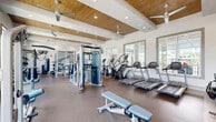 Located at 3200 Champion Ring Rd in Fort Myers, Florida, this fitness center offers a generously sized, single-floor layout designed to accommodate a comprehensive range of workout activities. The main workout area is anchored by high, beam-accented ceilings clad in warm wood paneling, complemented by recessed lighting and multiple ceiling fans that maintain a comfortable, airy atmosphere. Large multi-pane windows line the walls, flooding the space with abundant natural light and providing views of the surrounding outdoor greenery. The flooring throughout the main area features durable, rubberized material suited for heavy equipment use. The main floor is outfitted with an extensive selection of equipment, including multiple treadmills, elliptical machines, cable and pulley systems, free weights, barbells with plate racks, a punching bag, and Matrix-branded adjustable benches. Full-length mirrors extend along one wall, enhancing the sense of space. A separate studio room is accessible via glass-paneled partition walls and doors, creating a bright, open feel between the two areas. This studio features large windows, smooth light-toned flooring, a dramatic abstract wall mural, a large statement ceiling fan, spin bikes, stability balls, and a mounted exercise mat — ideal for group fitness or focused training sessions.