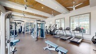 Located at 3200 Champion Ring Rd in Fort Myers, Florida, this fitness center offers a generously sized, single-floor layout designed to accommodate a comprehensive range of workout activities. The main workout area is anchored by high, beam-accented ceilings clad in warm wood paneling, complemented by recessed lighting and multiple ceiling fans that maintain a comfortable, airy atmosphere. Large multi-pane windows line the walls, flooding the space with abundant natural light and providing views of the surrounding outdoor greenery. The flooring throughout the main area features durable, rubberized material suited for heavy equipment use. The main floor is outfitted with an extensive selection of equipment, including multiple treadmills, elliptical machines, cable and pulley systems, free weights, barbells with plate racks, a punching bag, and Matrix-branded adjustable benches. Full-length mirrors extend along one wall, enhancing the sense of space. A separate studio room is accessible via glass-paneled partition walls and doors, creating a bright, open feel between the two areas. This studio features large windows, smooth light-toned flooring, a dramatic abstract wall mural, a large statement ceiling fan, spin bikes, stability balls, and a mounted exercise mat — ideal for group fitness or focused training sessions.