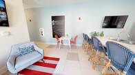 The 4830 Wescott Apartments clubhouse in Summerville, South Carolina is a thoughtfully designed single-floor community space that seamlessly blends functionality with a vibrant, coastal-inspired aesthetic. The interior is finished with large white tile flooring accented by decorative inset patterns, complemented by soft blue and warm beige walls that create a light and welcoming atmosphere throughout.

The open-concept layout connects a well-appointed community kitchen to a comfortable lounge and social area. The kitchen is outfitted with navy blue lower cabinetry paired with white upper cabinets featuring glass-front doors, stainless steel appliances including a French door refrigerator, double wall oven, and dishwasher, along with a spacious white quartz island with bar seating. A wall-mounted TV is positioned above for added convenience.

The lounge area offers flexible seating arrangements, including plush gray accent chairs arranged on a bold red-and-white striped area rug, bistro-style tables with red and navy woven chairs, and a dedicated community chalkboard wall branded with the 4830 Wescott Apartments identity. Natural light pours in through multiple windows, and a set of large glass double doors opens directly to the outdoor pool area, creating an effortless indoor-outdoor connection.