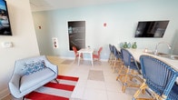 The 4830 Wescott Apartments clubhouse in Summerville, South Carolina is a thoughtfully designed single-floor community space that seamlessly blends functionality with a vibrant, coastal-inspired aesthetic. The interior is finished with large white tile flooring accented by decorative inset patterns, complemented by soft blue and warm beige walls that create a light and welcoming atmosphere throughout.

The open-concept layout connects a well-appointed community kitchen to a comfortable lounge and social area. The kitchen is outfitted with navy blue lower cabinetry paired with white upper cabinets featuring glass-front doors, stainless steel appliances including a French door refrigerator, double wall oven, and dishwasher, along with a spacious white quartz island with bar seating. A wall-mounted TV is positioned above for added convenience.

The lounge area offers flexible seating arrangements, including plush gray accent chairs arranged on a bold red-and-white striped area rug, bistro-style tables with red and navy woven chairs, and a dedicated community chalkboard wall branded with the 4830 Wescott Apartments identity. Natural light pours in through multiple windows, and a set of large glass double doors opens directly to the outdoor pool area, creating an effortless indoor-outdoor connection.