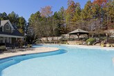 This inviting outdoor amenity area is thoughtfully designed to offer residents a comprehensive recreational retreat. At its centerpiece sits a generously sized swimming pool with clear, blue water, surrounded by a broad paver deck that provides ample room for lounging and socializing. The pool area is framed by mature trees and carefully maintained landscaping, creating a natural, park-like atmosphere.

Adjacent to the pool, a spacious paver patio features multiple seating arrangements, including wrought iron lounge chairs, dining tables, and a classic outdoor umbrella, offering comfortable spaces for relaxation and outdoor dining. A wood-framed gazebo with a shingled roof anchors one end of the patio, providing a shaded gathering spot that complements the overall design.

The surrounding landscape includes neatly trimmed hedgerows, ornamental plantings, and a stacked stone retaining wall that adds a refined architectural element to the grounds. Lamp posts and accent lighting suggest the space is designed for enjoyment both day and evening.

The community clubhouse building, clad in green siding with white trim, stone accents, and arched windows, borders the patio area and adds a classic, residential charm to the space. Together, these elements create a cohesive, welcoming outdoor environment ideal for both active recreation and quiet relaxation.