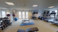This fitness center offers a comprehensive workout environment designed to accommodate a variety of exercise routines. The space is finished with striped carpeting underfoot and a textured ceiling fitted with recessed panel lighting, creating a clean and functional atmosphere. Natural light streams in through a set of French doors along one wall, which also provide access to an outdoor area, adding an open, airy feel to the room. The cardio section features an elliptical trainer and an additional cardio machine, ideal for endurance and low-impact workouts. A weight bench and adjustable equipment provide strength training options, while an array of stability and medicine balls in varying sizes supports balance, flexibility, and core conditioning exercises. Yoga mats laid out on the floor offer a dedicated area for stretching, floor work, or mindfulness routines. Decorative artwork on the walls adds a touch of visual interest to the otherwise utilitarian space. The layout is thoughtfully arranged to allow ample room for movement between equipment zones. Overall, this fitness center provides a well-rounded selection of tools to support cardiovascular fitness, strength training, and flexibility work in a comfortable, single-floor setting.