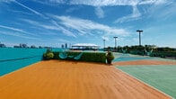 This dynamic rooftop venue offers a one-of-a-kind outdoor experience defined by bold color and an energetic atmosphere. The centerpiece is a generously sized multi-sport court finished in a striking palette of teal, orange, and green, suitable for pickleball, basketball, and other activities. Court markings are clearly delineated, and tall pole lighting ensures usability well into the evening hours. A covered event tent anchors one section of the rooftop, providing shade over a comfortable lounge area furnished with vibrant orange seating, coffee tables, and string lights that add warmth to the space. Adjacent to the tent, rows of lounge chairs line the perimeter, offering relaxed sunbathing spots with panoramic city skyline views. Decorative green hedgewall panels and potted flowering plants add a touch of greenery throughout. A turf lawn area with casual seating provides an additional social zone, while hammock chairs offer a leisurely retreat. A painted green masonry structure serves as a visual anchor and houses utility access. The rooftop perimeter features low walls and metal fencing, framing unobstructed views of the surrounding urban skyline on all sides. The overall ambiance is lively, colorful, and well-suited for social gatherings, athletic play, and events.