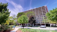 This space is situated within a striking multi-story high-rise building that showcases a classic mid-century architectural design. The building's exterior features a structured grid of white concrete columns and beams contrasting with warm brick tones, creating a bold and organized façade. Rows of private balconies span each floor, offering residents access to outdoor space with views of the surrounding greenery.

The building is framed by a lush landscape of mature deciduous trees, including a notable ornamental tree with deep burgundy foliage, contributing to the property's well-established character. The grounds are thoughtfully maintained, with manicured shrubs, mulched beds, and open green lawn areas enhancing the curb appeal.

The main entrance at ground level features a covered, columned entry with glass doors, providing a welcoming and accessible point of arrival. Ample surface parking is available along the front of the building. The overall setting conveys a sense of established, community-oriented living within a well-kept, park-like environment, offering both convenience and a connection to the natural surroundings.