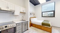 This thoughtfully designed studio in San Francisco makes excellent use of its compact footprint, offering a seamless blend of modern aesthetics and functional living. Light-toned hardwood-style flooring runs throughout, anchoring the clean, minimalist interior.

The open living area features soaring ceilings with exposed dark beams, white walls, and large windows that invite natural light and outdoor views. A wall-mounted TV, gray sofa, and nesting coffee tables create a comfortable and practical lounge space, complemented by a wall-mounted mini-split unit for climate control.

The kitchen is well-appointed with two-tone cabinetry — white upper cabinets and gray lower drawers — paired with a quartz countertop, stainless steel sink, electric range, and full-size refrigerator. A range hood and ample storage make this kitchen highly functional despite the compact layout.

The sleeping nook is efficiently arranged with a platform bed featuring built-in storage drawers beneath, framed art on the wall, and a window overlooking the outdoors. The space feels private and restful.

The bathroom maintains the studio's clean design language, with light flooring, a vanity with cabinetry, and a walk-in shower area, all finished in crisp white tones with black hardware accents. Every element of this studio is optimized for modern urban living.
