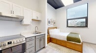This thoughtfully designed studio in San Francisco makes excellent use of its compact footprint, offering a seamless blend of modern aesthetics and functional living. Light-toned hardwood-style flooring runs throughout, anchoring the clean, minimalist interior.

The open living area features soaring ceilings with exposed dark beams, white walls, and large windows that invite natural light and outdoor views. A wall-mounted TV, gray sofa, and nesting coffee tables create a comfortable and practical lounge space, complemented by a wall-mounted mini-split unit for climate control.

The kitchen is well-appointed with two-tone cabinetry — white upper cabinets and gray lower drawers — paired with a quartz countertop, stainless steel sink, electric range, and full-size refrigerator. A range hood and ample storage make this kitchen highly functional despite the compact layout.

The sleeping nook is efficiently arranged with a platform bed featuring built-in storage drawers beneath, framed art on the wall, and a window overlooking the outdoors. The space feels private and restful.

The bathroom maintains the studio's clean design language, with light flooring, a vanity with cabinetry, and a walk-in shower area, all finished in crisp white tones with black hardware accents. Every element of this studio is optimized for modern urban living.