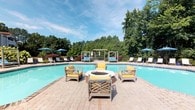 This single-story clubhouse offers a well-appointed amenity space designed for both relaxation and recreation. The centerpiece is a generously sized outdoor swimming pool with clear turquoise water, surrounded by a brick-paver pool deck. Poolside seating includes lounge chairs with blue umbrellas and a dedicated pergola structure at the far end, providing shaded gathering areas. A circular fire pit with lattice-style wooden chairs cushioned in yellow anchors the deck as a social focal point, with lush mature trees forming a natural backdrop.

Inside, the recreation room features vaulted ceilings with a skylight that floods the space with natural light. Light hardwood flooring runs throughout, complementing the warm yellow walls. A full-size pool table is prominently positioned beneath the soaring roofline, while a workout area with treadmills is visible through a frosted interior window. A lounge area with teal upholstered seating and a workstation provide additional functionality.

Large picture windows along one wall frame unobstructed views of the pool and patio. French doors open to a covered outdoor patio equipped with a ceiling fan and teak seating, bridging the indoor and outdoor spaces seamlessly. The overall ambiance is bright, welcoming, and resort-inspired.