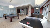 This contemporary amenity lounge is thoughtfully designed to accommodate both active recreation and relaxed socializing on a single level. The space is anchored by a full-size ping pong table and a dark-wood pool table, making it well-suited for recreational use. Light-toned tile flooring runs throughout, contributing to a clean, modern aesthetic, while recessed ceiling lighting and pendant fixtures provide consistent, well-distributed illumination across the room.

A casual dining area with bar-height tables and metal stools occupies the mid-section of the lounge, complemented by a wall-mounted flat-screen television — ideal for social gatherings or watching sports. Toward the rear, a built-in linear fireplace framed by stacked stone tile wall accents adds a warm, upscale element to the space. Additional seating is arranged near the fireplace, creating a comfortable lounge area. Frosted glass partitions offer subtle spatial division without compromising the open feel of the room. The overall design blends industrial and contemporary influences, resulting in a polished, multifunctional common space.