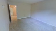 This two-floor space presents a straightforward and functional interior design with a neutral color palette throughout. The main living area visible in the image features wall-to-wall carpeting in a light beige tone, complemented by smooth, cream-colored walls and clean white baseboards that contribute to a bright and understated aesthetic. Multiple electrical outlets are positioned along the walls, offering practical convenience for furniture arrangement and everyday use. An open doorway connects the carpeted living area to an adjacent room finished with tile flooring, which is illuminated by a ceiling-mounted light fixture. This transitional space, possibly an entryway or dining area, features what appears to be a door with vertical blinds, suggesting access to the outdoors or another section of the unit. The overall ambiance is clean and move-in ready, with a simple, unfussy design that provides a neutral canvas for personalization. The two-floor layout offers additional living space beyond what is immediately visible, adding versatility to the overall footprint of the home.