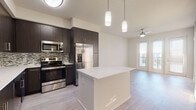 This contemporary single-floor residence showcases a thoughtfully designed open-concept layout that seamlessly connects the kitchen and living areas. The kitchen is a standout feature, boasting dark espresso cabinetry that extends to the ceiling, a multicolored mosaic tile backsplash, crisp white countertops, and a full suite of stainless steel appliances including a French door refrigerator with ice maker, electric range, built-in microwave, and dishwasher. A central island provides additional prep space and casual seating. Pendant lighting above the island adds a stylish touch to the space. The living area is bathed in natural light streaming through large floor-to-ceiling windows and a glass door that opens to a balcony, complemented by a ceiling fan overhead. Wide-plank light wood-style flooring runs consistently throughout the main living spaces, lending warmth and visual continuity. The bedroom is generously proportioned with a bright window that invites natural light. The bathroom is finished with a dark vanity, white countertop, large-format tile flooring, and a spacious walk-in glass-enclosed shower. Overall, the space presents a clean, modern aesthetic with high-quality finishes throughout.