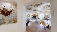 This thoughtfully designed single-floor space offers a seamless blend of classical architectural detail and contemporary comfort across its well-appointed rooms. The most striking architectural feature is the elaborately detailed coffered ceiling in the main living area, adorned with ornate moldings and decorative medallions that lend a distinctly grand character to the space.

The primary living room centers around a natural stone fireplace with a handcrafted wooden mantle, flanked by built-in wall niches ideal for display. Light wood-look flooring runs throughout, complementing the warm, neutral color palette. Arched passageways connect the rooms, reinforcing the classical aesthetic. A secondary living area, framed by large arched windows with vertical blinds, allows ample natural light and accommodates a workspace.

The dining area features a curved architectural divider that creates an intimate setting, with arched windows providing views to the exterior. A separate seating nook with decorative columns and recessed arched alcoves adds a refined touch. Throughout the space, wall sconces provide warm ambient lighting, enhancing the overall inviting and polished atmosphere.