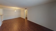 This single-floor residential unit offers a practical and well-organized layout designed for comfortable everyday living. The entry opens into a spacious living area finished with rich, dark hardwood-style flooring that extends throughout the main living spaces, creating a warm and cohesive aesthetic. A half-wall partition separates the entry from the adjoining area, and a pendant light fixture adds a touch of character to the dining space. The kitchen is efficiently designed with white cabinetry featuring modern hardware, light-toned countertops, and a black electric range with an overhead ventilation hood, providing a clean contrast against the bright cabinetry. The bedroom features wall-to-wall carpeting in a neutral tone, a window dressed with horizontal blinds that allows natural light to filter in, and a textured ceiling that carries throughout the unit. The bathroom is finished with light ceramic tile flooring, a white vanity with stone-look countertops, and a full tub-shower combination surrounded by classic white subway tile. Neutral gray walls throughout the unit provide a versatile backdrop, and the overall layout, as seen from the dollhouse view, reflects a compact yet thoughtfully arranged floor plan suited for a range of living arrangements.