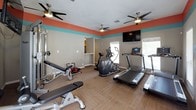 This fitness center is a fully equipped exercise space designed to support a well-rounded workout routine. The room is immediately recognizable by its bold and distinctive color scheme — a coral-orange upper wall band paired with a teal stripe, set against neutral gray lower walls, creating a vibrant and energizing atmosphere. Durable carpet tile flooring covers the space throughout, providing cushioning underfoot during workouts.

The cardio section is well-stocked with multiple treadmills, an Octane elliptical cross-trainer, and a stationary bike, offering a variety of aerobic exercise options. Wall-mounted flat-screen televisions are positioned for easy viewing during cardio sessions. The strength training area includes a cable machine, adjustable weight bench, additional free-standing benches, and a compact ab trainer, catering to a range of resistance and toning exercises.

A standout architectural feature is the stone-surround fireplace, adding an unexpected and welcoming element to the space. Multiple windows fitted with white blinds allow natural light to filter in from multiple directions. Ceiling fans are distributed throughout, ensuring comfortable air circulation. A set of French doors provides access to the exterior, further enhancing the room's openness.