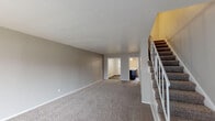 This two-story residence is thoughtfully laid out across two levels, offering a functional and comfortable living environment. The main level features a spacious living area with neutral-toned carpet, light gray walls, and white trim throughout. A front entry with a white door and large vertical blinds allows natural light to filter in, while a carpeted staircase with white balusters provides access to the upper floor. An adjacent room on the main level opens to additional living space, also carpeted and bathed in neutral tones.

The kitchen is a standout feature, equipped with dark cabinetry contrasted by white countertops and white appliances, including a coil-burner range and refrigerator. A peninsula with a breakfast bar separates the kitchen from the adjoining area, enhancing functionality. Dark wood-style flooring runs throughout the kitchen and extends into the bathrooms. A window above the sink brings in natural light.

Upstairs, multiple carpeted bedrooms each include ceiling fans with light fixtures and windows dressed with blinds or vertical blinds. One bedroom opens directly to the staircase landing. The bathroom features a tub-shower combination with white tile surround, a dark vanity, and wood-style flooring, completing the cohesive design found throughout the home.