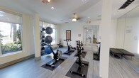 This single-floor fitness space is designed to support a well-rounded workout routine in a clean, contemporary setting. The room features light gray wood-look flooring throughout, providing a neutral and polished foundation for the exercise equipment. A row of stationary spin bikes positioned on protective rubber mats forms the centerpiece of the space, offering dedicated cycling stations for cardiovascular training.

Along one wall, a vertical rack displays an assortment of medicine balls and exercise balls in varying sizes, providing easy access to strength and flexibility training tools. Large windows allow generous natural light to fill the room, creating a bright and energizing atmosphere conducive to physical activity. The white walls and recessed ceiling lights further contribute to the open, airy feel of the space.

A ceiling fan provides additional air circulation, enhancing comfort during intense workouts. Glass-paneled double doors offer a clear view into an adjacent area, suggesting connectivity to broader amenity spaces. The overall layout is thoughtfully organized, balancing equipment accessibility with open floor space for stretching or mat-based exercises.