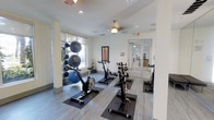 This single-floor fitness space is designed to support a well-rounded workout routine in a clean, contemporary setting. The room features light gray wood-look flooring throughout, providing a neutral and polished foundation for the exercise equipment. A row of stationary spin bikes positioned on protective rubber mats forms the centerpiece of the space, offering dedicated cycling stations for cardiovascular training.

Along one wall, a vertical rack displays an assortment of medicine balls and exercise balls in varying sizes, providing easy access to strength and flexibility training tools. Large windows allow generous natural light to fill the room, creating a bright and energizing atmosphere conducive to physical activity. The white walls and recessed ceiling lights further contribute to the open, airy feel of the space.

A ceiling fan provides additional air circulation, enhancing comfort during intense workouts. Glass-paneled double doors offer a clear view into an adjacent area, suggesting connectivity to broader amenity spaces. The overall layout is thoughtfully organized, balancing equipment accessibility with open floor space for stretching or mat-based exercises.