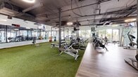 Located at 1800 N Stone Ave in Tucson, Arizona, this single-floor fitness facility offers a spacious and thoughtfully designed exercise environment spanning a generous footprint. The space is divided into several distinct functional zones, each tailored to a specific type of training.

The strength and functional training area features artificial turf flooring, weight benches, power racks, free weights, and medicine balls, all positioned beneath an exposed ceiling with industrial-style ductwork and circular halo lighting fixtures. Full-length mirrors line the walls, enhancing the sense of openness. Mounted televisions provide visual engagement throughout this zone.

Transitioning to the hardwood-style flooring section, the cardio zone is anchored by large windows that invite abundant natural light and views of the surrounding Tucson streetscape. Curved treadmills, stair climbers, and stationary bikes are neatly arranged in this bright, airy area.

A dedicated pilates and stretching zone features reformer machines positioned near glass entry doors that further brighten the space with natural light. Adjacent to this area, a golf simulator bay with projection equipment and a hitting mat offers a unique recreational amenity.

Throughout the facility, the exposed ceiling with visible ductwork and structural columns contribute to an open, industrial-modern aesthetic, while varied flooring materials help define each zone clearly.