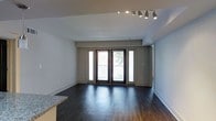 This contemporary single-floor space showcases a thoughtfully designed open-concept layout that seamlessly blends style and functionality. The living area is anchored by rich, dark hardwood flooring that extends throughout, creating a warm and sophisticated aesthetic against the crisp white walls and ceiling.

A standout feature of the main living space is the set of elegant French doors flanked by full-length sidelights, which flood the interior with natural light while providing direct access to a private balcony. The outdoor views and greenery visible through the glass add a refreshing sense of connection to the outdoors.

Overhead, a modern adjustable track lighting system provides versatile, directional illumination, making the space both practical and visually dynamic. The open layout creates a generous sense of space, with clean sightlines from the kitchen area — hinted at by the granite countertop visible at the edge of the frame — through to the living area and beyond.

The neutral color palette of soft whites and grays throughout ensures a timeless, move-in-ready feel, while the high-quality finishes, including the stone countertop and hardwood floors, reflect a commitment to modern, upscale living. The overall ambiance is bright, airy, and polished.