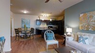 This single-story residence features an open-concept layout that seamlessly connects the kitchen, dining, and living areas into one cohesive, flowing space. Hardwood-style flooring runs throughout, lending warmth and continuity across each zone. The kitchen is outfitted with dark cabinetry, black appliances including a refrigerator, microwave, and range, and a dark stone countertop. A central island provides additional prep space and serves as a natural divider between the kitchen and the rest of the living area. Pendant lighting and a multi-bulb track light illuminate the kitchen, while a ceiling fan with a light fixture anchors the main living space above. The dining area accommodates a compact table with chairs and is accented by a bold coastal-inspired wall canvas, adding personality to the space. The living area is furnished with a light-toned sofa and an ornate accent chair featuring a teal cushion, contributing to the space's relaxed, coastal aesthetic. Neutral wall tones with a contrasting gray accent wall add depth, while the overall layout maximizes the sense of openness and functionality throughout.