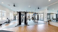 This single-level fitness center in Athens, Alabama, offers a generously sized, thoughtfully designed workout space suited for a full range of exercise routines. The interior combines hardwood-style flooring in the open stretching and free-weight areas with durable rubber matting beneath the strength and cardio equipment, providing both visual warmth and functional performance underfoot.

The cardio zone is well-appointed with multiple Bodycraft treadmills, elliptical trainers, a stair climber, and stationary bikes, all positioned along large, multi-pane windows that flood the space with natural light and offer exterior views of the surrounding community. Wall-mounted flat-screen televisions are positioned throughout for entertainment during workouts.

The strength training area features a TKO power rack, a cable crossover machine, TRX suspension trainer, a heavy bag, and a dedicated free-weight station with an adjustable bench and dumbbell rack. A full-length mirror wall enhances the sense of openness and allows users to monitor form.

Recessed lighting and ceiling fans maintain a comfortable, well-lit environment. A distinctive glass roll-up garage door connects to an adjacent outdoor area, promoting airflow and an open-air feel. The entrance features a bold charcoal accent wall with vertical wood slat detailing, framing stylish glass French doors and adding a modern, polished aesthetic to the overall design.