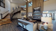 This two-story amenity space blends rustic warmth with modern sophistication through a carefully curated design palette. The centerpiece is a grand, double-height lobby lounge anchored by stacked stone columns, rich wood-paneled accent walls, and a sleek linear gas fireplace framed in stone. Soaring ceilings draw the eye upward to a mezzanine level accessible by a striking open-riser staircase with industrial metal railings. Generous seating arrangements, dark hardwood-style flooring, and curated wall art create an inviting, lodge-inspired atmosphere.

The bar and lounge area features vaulted ceilings with exposed wood beams, pendant globe lighting, and a full bar with quartz countertops and bar stool seating. Large windows and glass doors flood the space with natural light and offer views of the surrounding exterior.

A fully equipped communal kitchen includes a large central island with a gas cooktop, stainless steel appliances, dark cabinetry, and a textured tile backsplash — ideal for group gatherings and events.

Additional spaces on the second floor include a cozy game room with wood-paneled walls, natural light from oversized windows, and a classic pinball machine, as well as intimate lounge nooks with plush seating for relaxed socializing.
