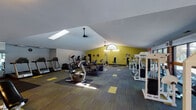 Located at 110 Presidio Ct in Schaumburg, IL, this single-floor fitness center offers a comprehensive workout environment designed to accommodate a wide range of exercise needs. The space is generously sized, with a vaulted ceiling on one end highlighted by a skylight that draws in natural light, creating an open and energizing atmosphere. Ceiling fans contribute to comfortable airflow throughout the facility.

The cardio zone is well-appointed with multiple treadmills, elliptical trainers, stair climbers, and stationary bikes, including recumbent models, all arranged on protective rubber matting. The strength training area features a variety of cable and weight machines, a dedicated dumbbell rack, and additional functional training stations positioned along the perimeter.

The flooring consists of multi-toned modular carpet tiles in blue, gray, and yellow, lending a modern, structured aesthetic to the space. Bold yellow accent walls add visual energy and contrast with the otherwise neutral palette.

Large floor-to-ceiling windows along one wall offer direct views of an outdoor pool and patio area, providing natural light and a seamless visual connection to the exterior amenity space. Brick column accents frame the window wall, adding architectural character to the overall design.