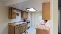 This single-floor space centers around a galley-style kitchen that showcases a practical and efficient layout. The kitchen features warm-toned wood cabinetry both above and below the countertops, providing generous storage throughout. Upper cabinets line one wall, offering ample space for pantry essentials, while lower cabinets and drawers maximize organizational potential at counter level.

The countertops are finished in a light wood butcher-block style, lending a natural, warm aesthetic to the cooking space. A built-in dishwasher and a white refrigerator are seamlessly integrated into the layout, while a white gas cooktop sits on the opposing counter, making the kitchen well-equipped for meal preparation. A single-basin sink is positioned along the main counter run, accompanied by a standard faucet.

A notable architectural feature is the recessed skylight panel in the ceiling, which introduces natural overhead light into the otherwise enclosed kitchen space. The light-colored walls and neutral flooring contribute to a bright, clean atmosphere. A doorway at the far end of the kitchen connects to an adjacent area, suggesting a logical and connected flow throughout the single-level layout.