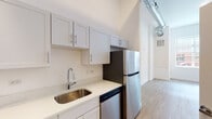 This single-floor unit presents a clean, modern aesthetic throughout, characterized by crisp white walls, light-toned hardwood-style flooring, and exposed industrial ductwork that adds a loft-like character to the space. The kitchen is efficiently designed with white shaker-style cabinetry, light quartz countertops, a stainless steel single-basin sink, and a full suite of stainless steel appliances including a refrigerator, range, built-in microwave, and dishwasher. The layout provides ample counter space and storage. The living area benefits from a large multi-pane window that invites generous natural light and offers a street-level view, creating an airy and welcoming atmosphere. The bedroom is a well-proportioned, light-filled room featuring tall ceilings and an open closet with a hanging rod, offering practical storage. The bathroom is simply appointed with a white vanity and integrated sink, a wall-mounted mirror with overhead lighting, a glass-enclosed shower, and wood-style flooring consistent with the rest of the unit. Overall, the space maintains a cohesive, minimalist design with industrial accents throughout.